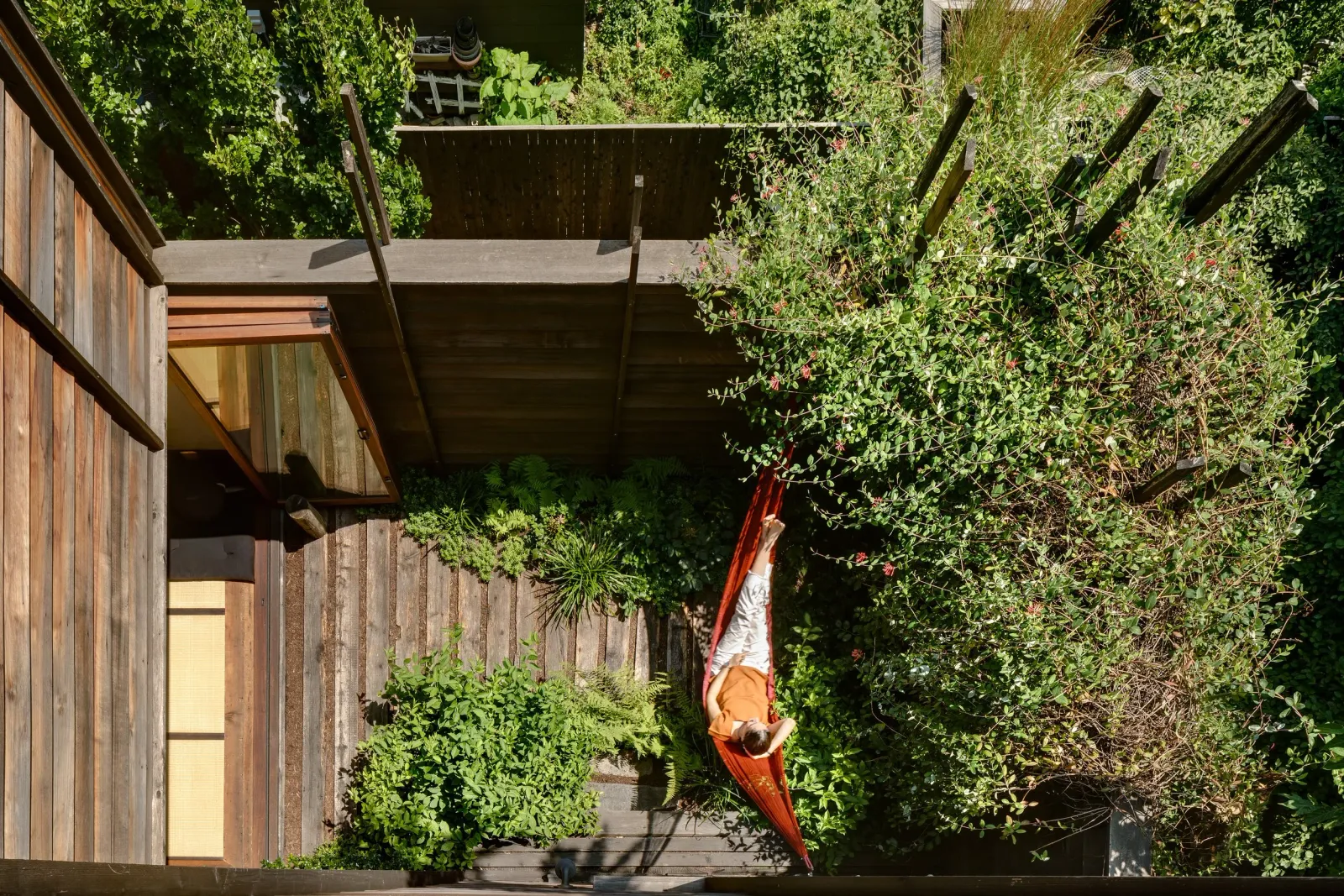 A woman lying on an orange hammock in a lush green backyard garden, surrounded by plants and trees with a wooden fence.