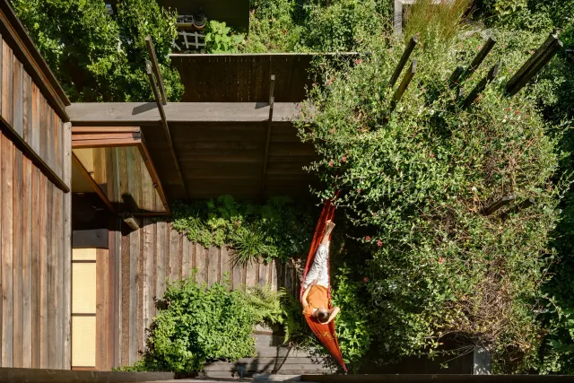 A woman lying on an orange hammock in a lush green backyard garden, surrounded by plants and trees with a wooden fence.