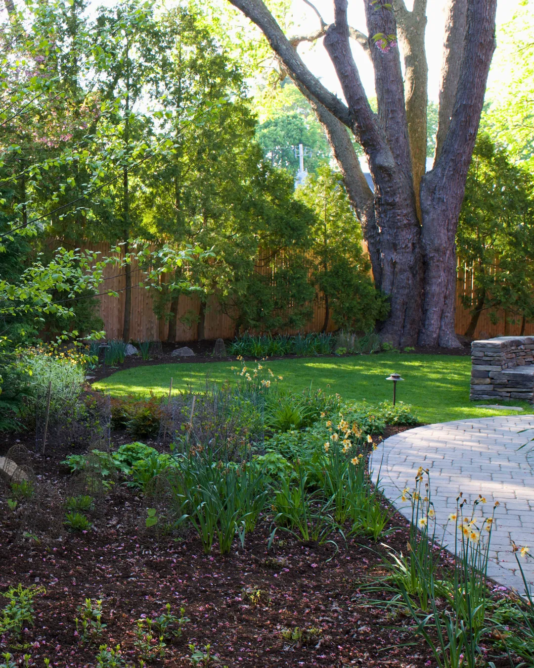 A serene garden with vibrant green grass, surrounded by trees and flowering plants. There is a brick pathway curving through the garden, leading to a cluster of large trees at the back. Sunlight filters through the trees, casting dappled shadows.