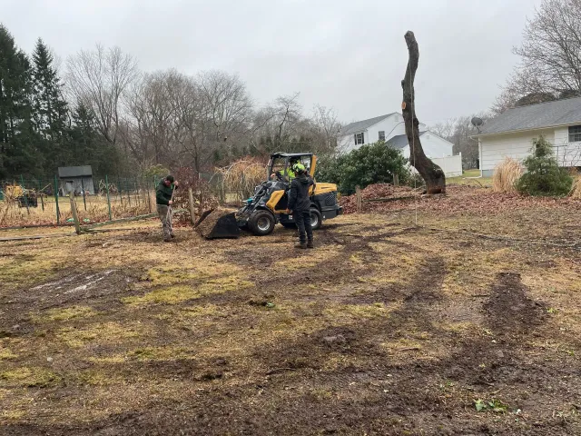 Workers with a yellow machine clearing a muddy, burned yard with trees and houses in the background