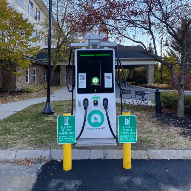 Electric vehicle charging station with two charging cables, surrounded by yellow safety bollards, in an outdoor parking area with trees, benches, and a building in the background.