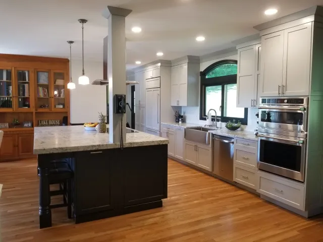 Modern kitchen with white cabinets, a black-framed window, stainless steel appliances, and a wooden floor. A kitchen island has a black base and a stone countertop. There are pendant lights hanging above the island and recessed ceiling lights. A built-in bookshelf with books and decorative items is visible in the background.