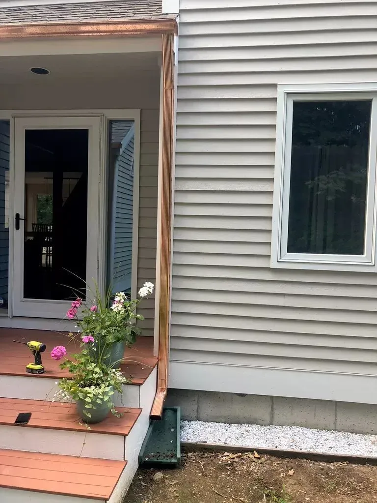 Front porch of a house with potted flowers, a black door, and beige siding beside a window.
