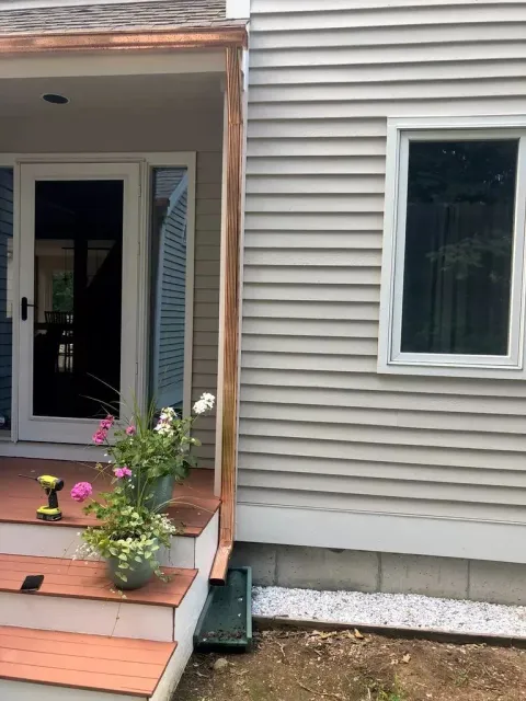 Front porch of a house with potted flowers, a black door, and beige siding beside a window.