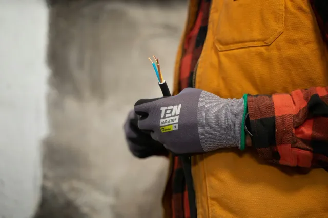 A person wearing a gray glove with the brand 'TEN' is holding a black cable with three exposed colored wires (blue, yellow, brown). The person is wearing an orange work vest over a red and black plaid shirt.