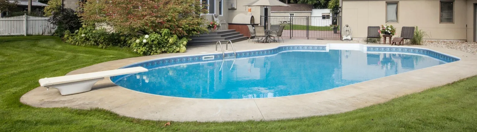 Backyard with in-ground swimming pool, diving board, lawn, patio furniture, and surrounding greenery.