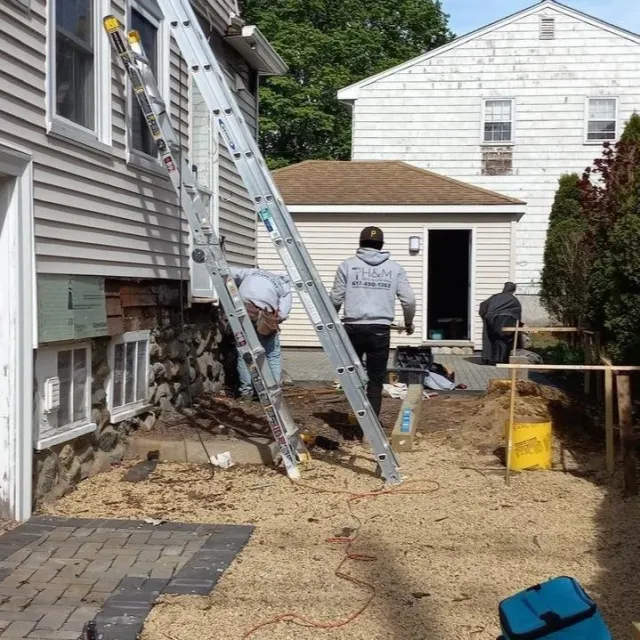 Workers repairing or inspecting the exterior of a house, with a ladder leaning against the house, and construction materials and tools around them in a backyard.
