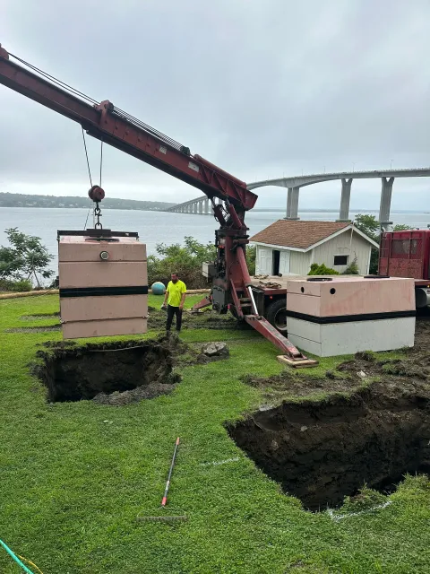 Crane lifting a pink utility box near sinkholes on a grassy site, workers nearby and a bridge in the background