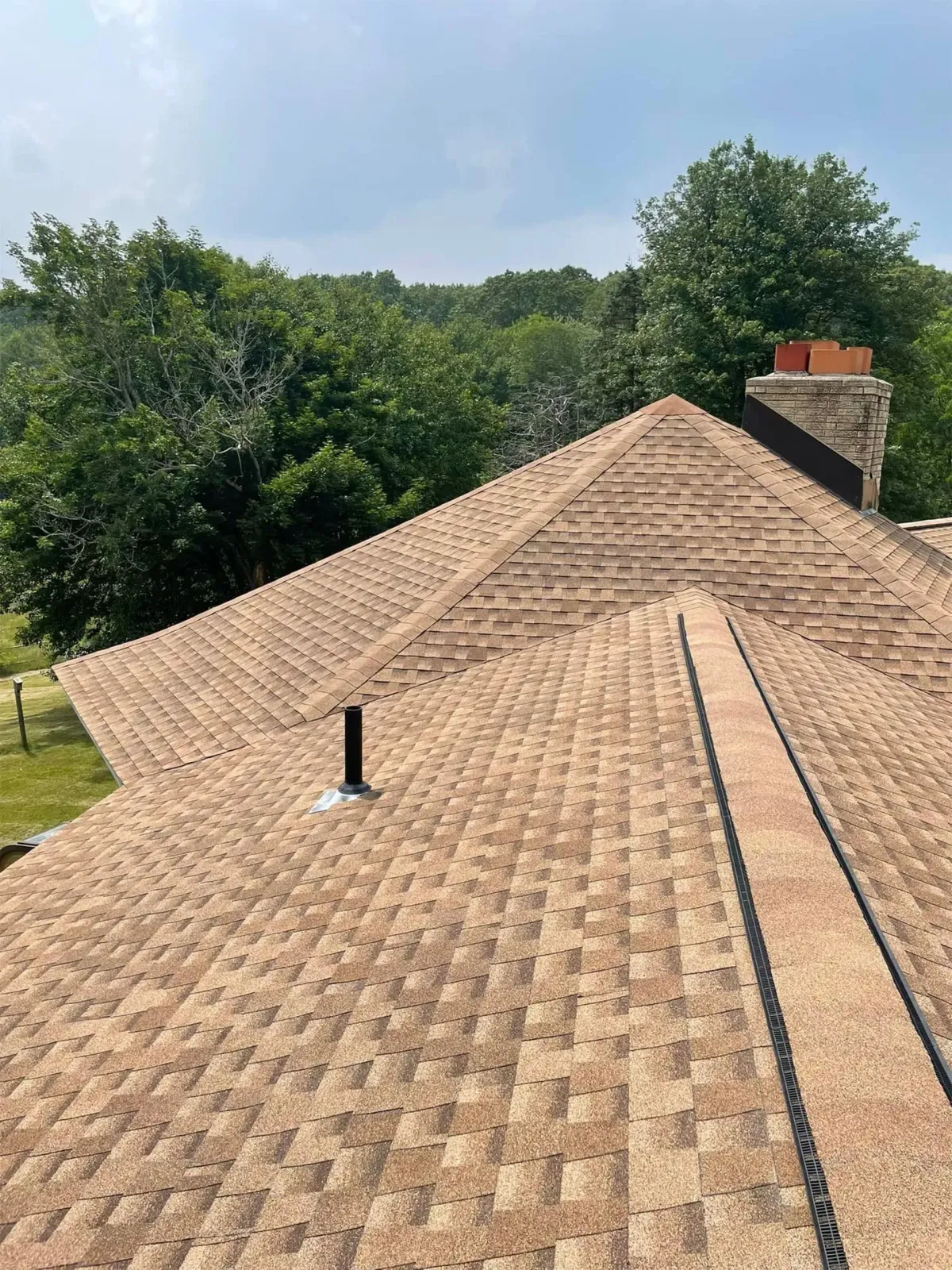 Tan shingle roof with a chimney and vent, surrounded by green trees under a blue sky
