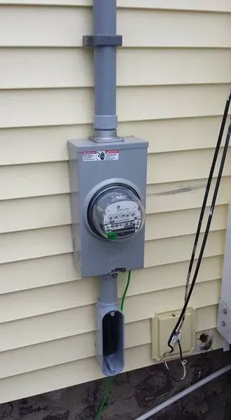 Gray electrical meter box on a yellow house siding, with conduit and a grounded green wire.