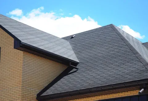 Brick house roof with dark gray shingles against a blue sky with clouds