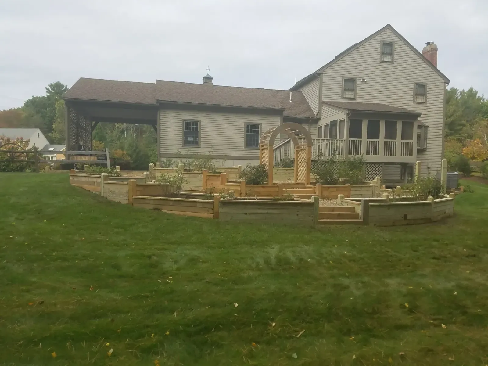 Rustic farmhouse with a wooden deck and fenced garden in a grassy yard under an overcast sky