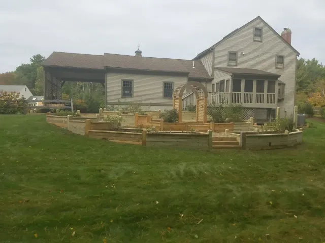 Rustic farmhouse with a wooden deck and fenced garden in a grassy yard under an overcast sky