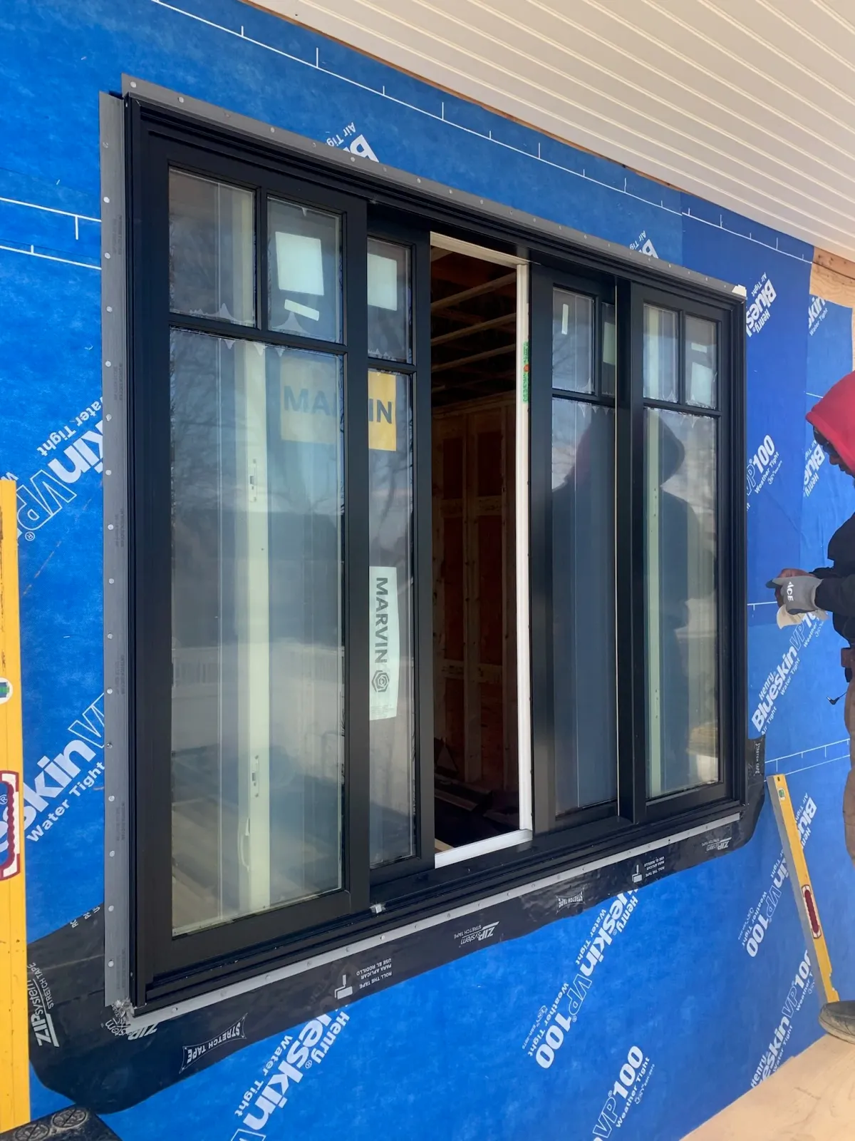 A black sliding glass door installed on a building under construction, with blue weatherproofing material and a person in a red hoodie working nearby. Scituate, Norwell, Hanover, Hanson, Marshfield, South Shore. Massachusetts Local Construction.