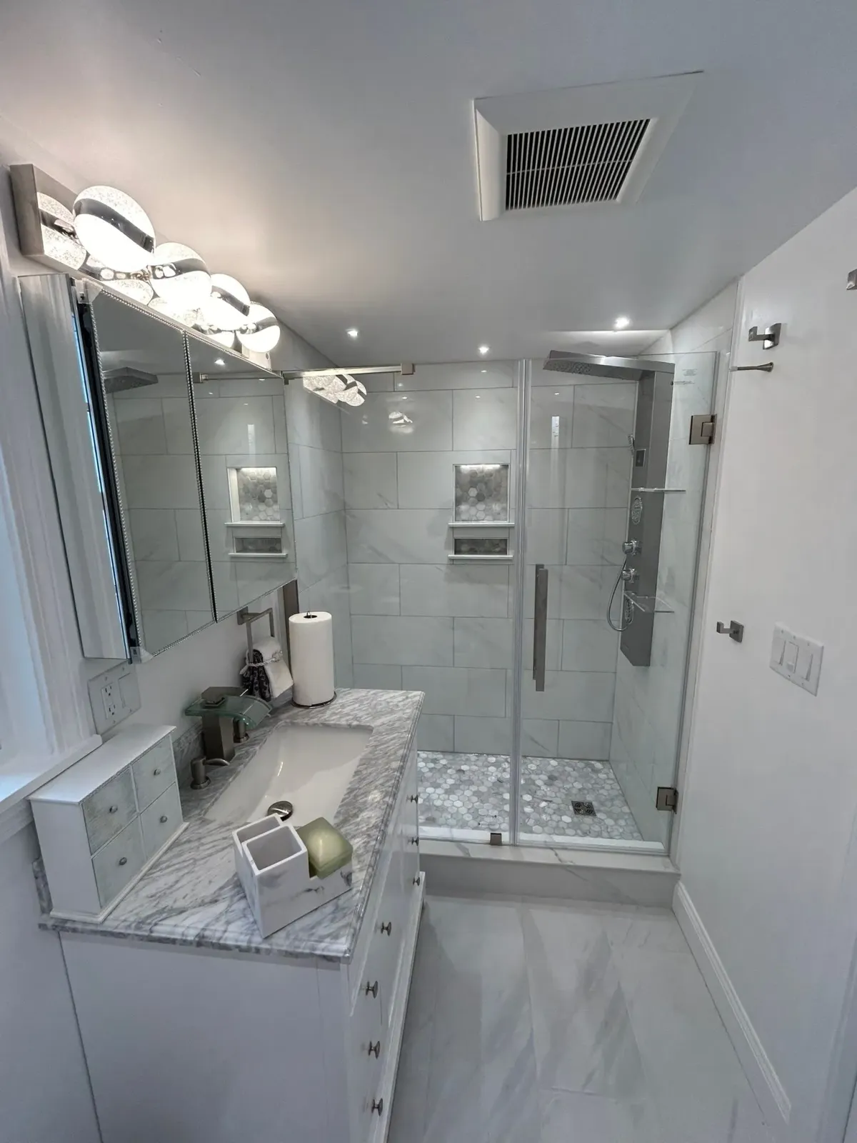 Modern bathroom with white and gray tile, glass shower, vanity, and overhead lighting.