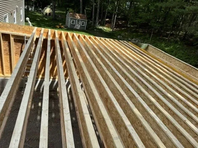 Wooden roof framing under construction on a house, with exposed rafters and a wooded background.
