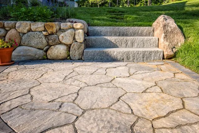 Stone patio with rock wall and granite steps leading to a grassy area, with a potted plant on the left.