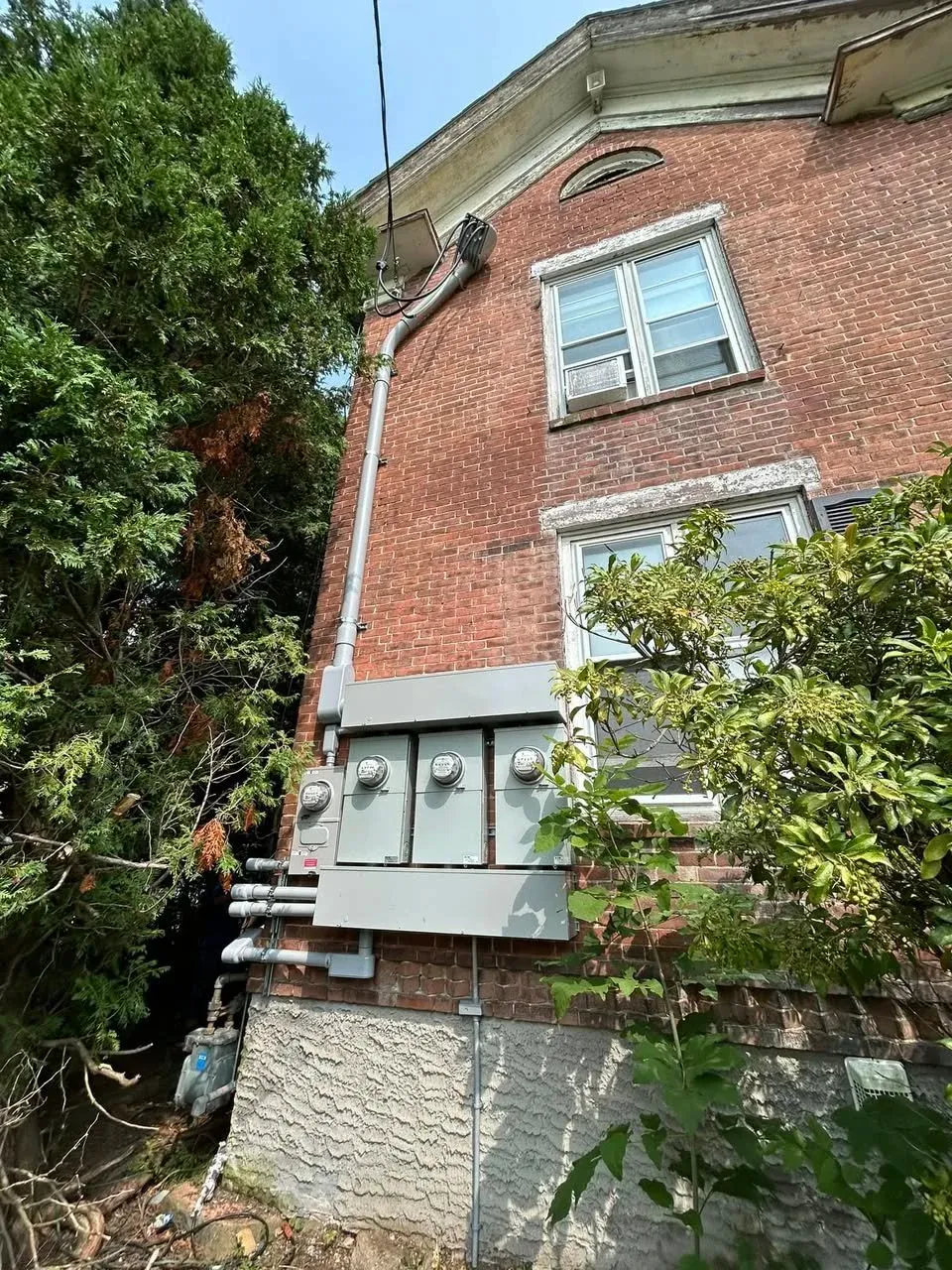Side of a red brick building with utility meters and electrical conduit, surrounded by greenery.