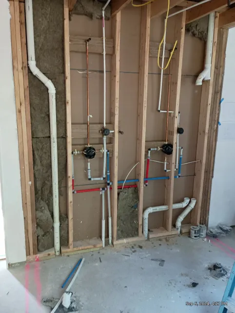 Open wall with plumbing pipes and water shutoff valves installed, framing in place, construction materials on floor.