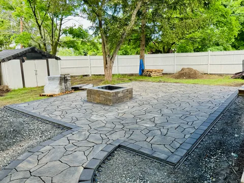 A newly installed stone patio stretches across a backyard, featuring irregularly shaped paving stones in varying shades of gray. A square fire pit sits at the center of the patio, while gravel borders and curved pathways frame the space. Mature trees and a white privacy fence surround the yard, creating an open outdoor gathering area beside the house.