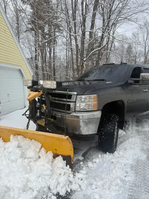 Black pickup truck clearing snow with front plow in a snowy driveway by a yellow house