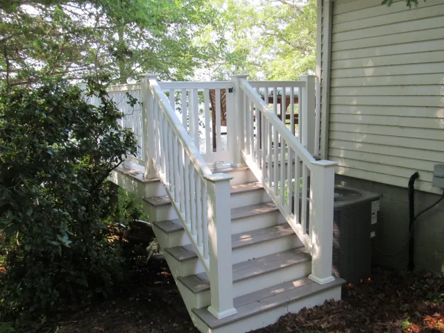 Outdoor white wooden staircase with railing leading to a deck, surrounded by green foliage, attached to a house.