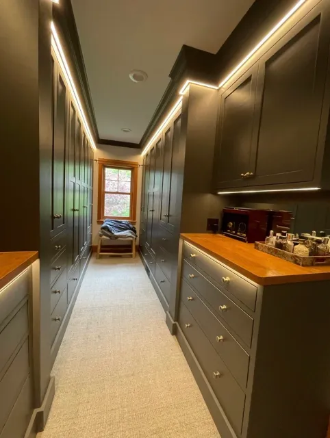 A narrow walk-in closet with dark gray built-in cabinets and drawers on both sides, a beige carpeted floor, and a window at the end with a small bench beside it holding clothes. The ceiling has recessed lighting with LED strip accents.