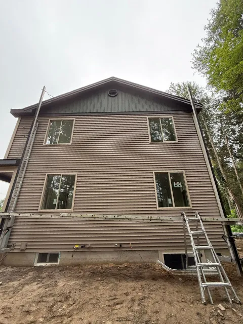 Weathered wood-sided building with windows. Ladder leaned against it, cloudy sky overhead.