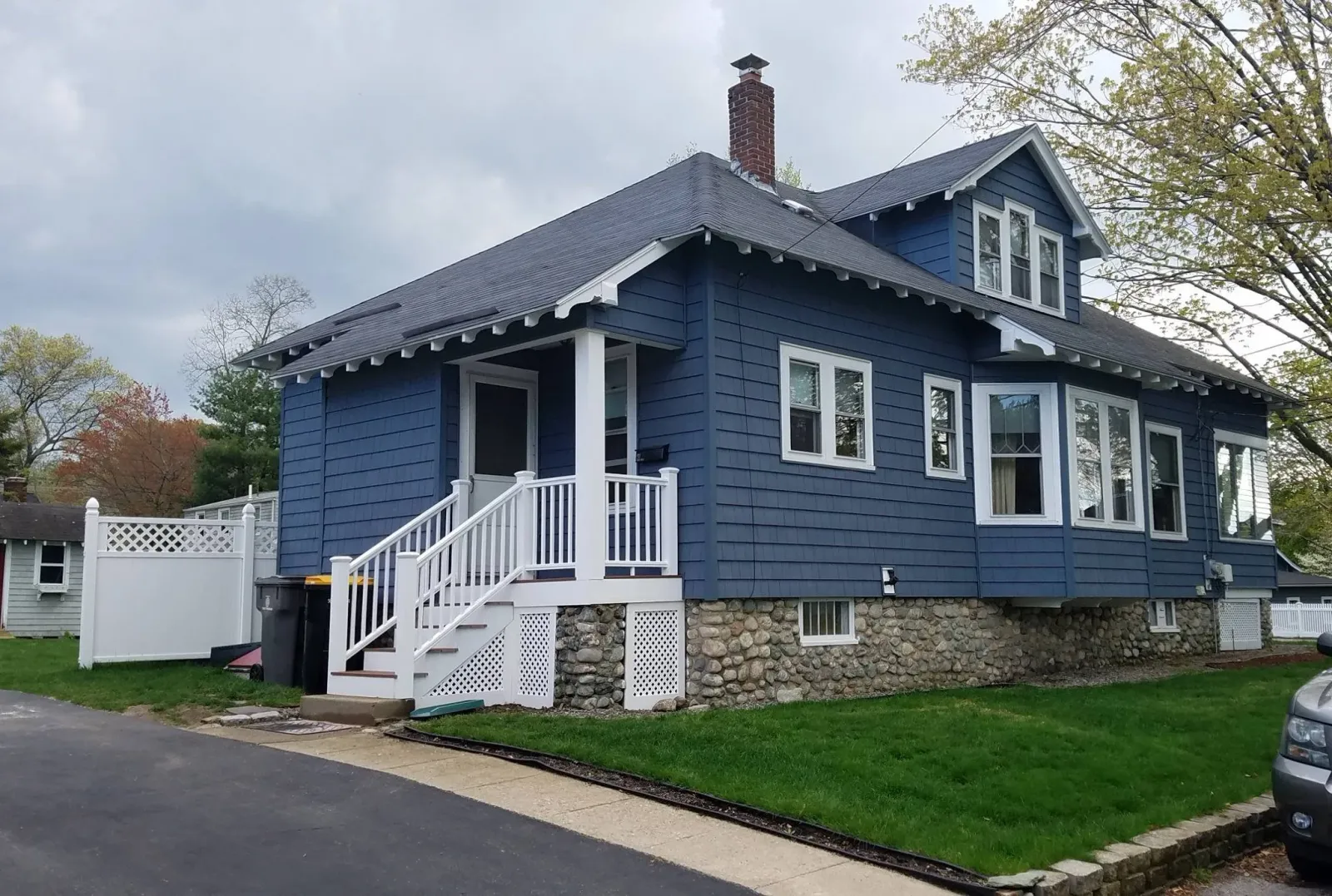 A blue two-story house with white trim, a stone foundation, a small front porch with stairs, and a chimney, surrounded by trees and a grassy yard.