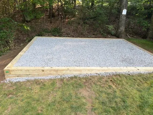 Gravel-filled rectangular base, likely for a shed, framed by wooden beams on a grassy lawn. Forest in the background.