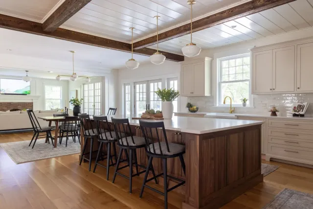 A bright kitchen with a wooden island and black bar stools, featuring white cabinetry, pendant lights, and wood floors.