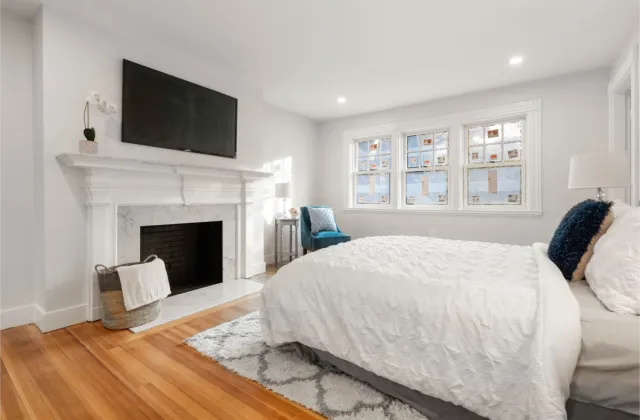 Bright bedroom with a white bed, fireplace, TV, and chair by the window showcases expert design.
