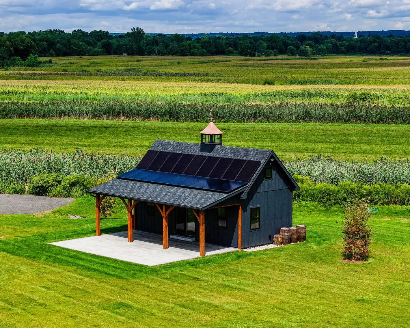 Small dark shed with solar panels in a green field, surrounded by farmland under a cloudy sky