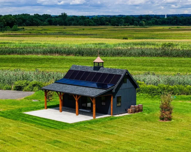 Small dark shed with solar panels in a green field, surrounded by farmland under a cloudy sky