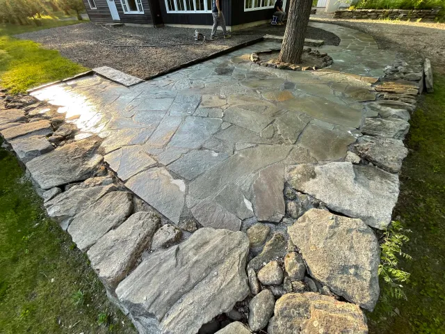 A newly constructed stone patio with large flat stones and a border of larger, rougher stones surrounds a tree in a backyard. A person is working nearby, and a house is visible in the background.
