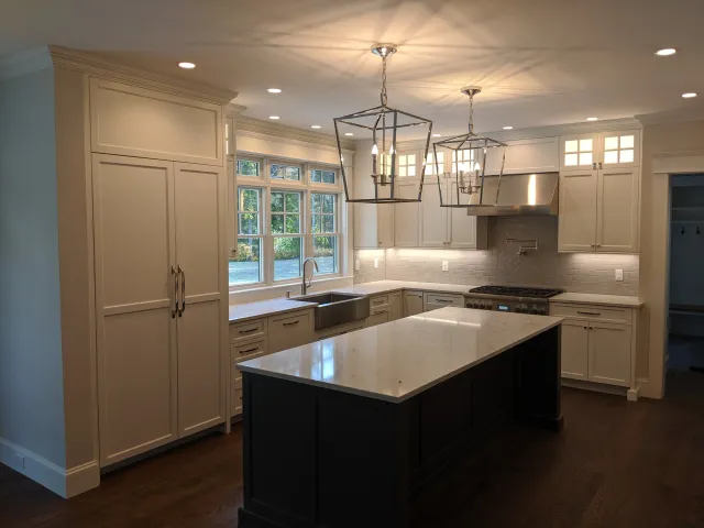 Modern kitchen with white cabinets, large window, central island with dark base and white marble top, stainless steel appliances, pendant lights, and hardwood floors.