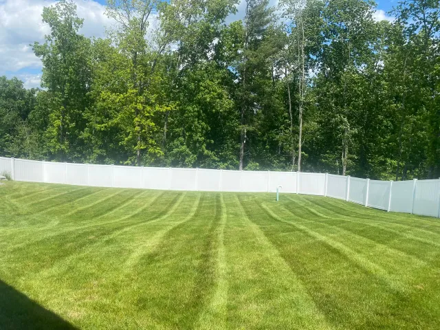 A freshly mown lawn with striped patterns, enclosed by a white vinyl fence in front of a line of green trees.