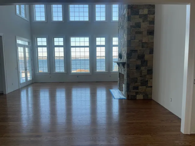 Spacious room with hardwood floors, large windows overlooking water, and a stone fireplace.