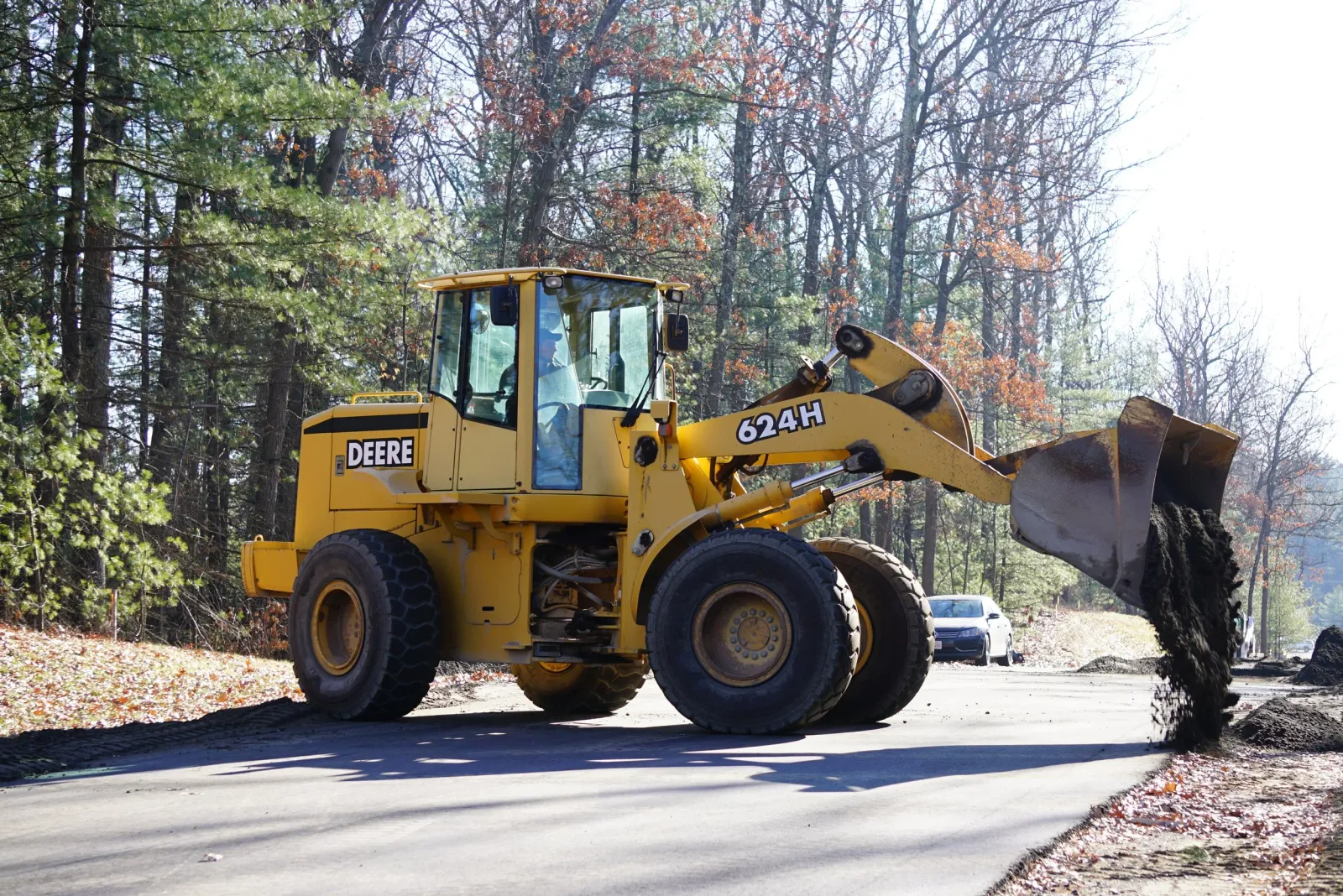 A yellow John Deere front loader bulldozer moving dirt in a wooded area during daytime.