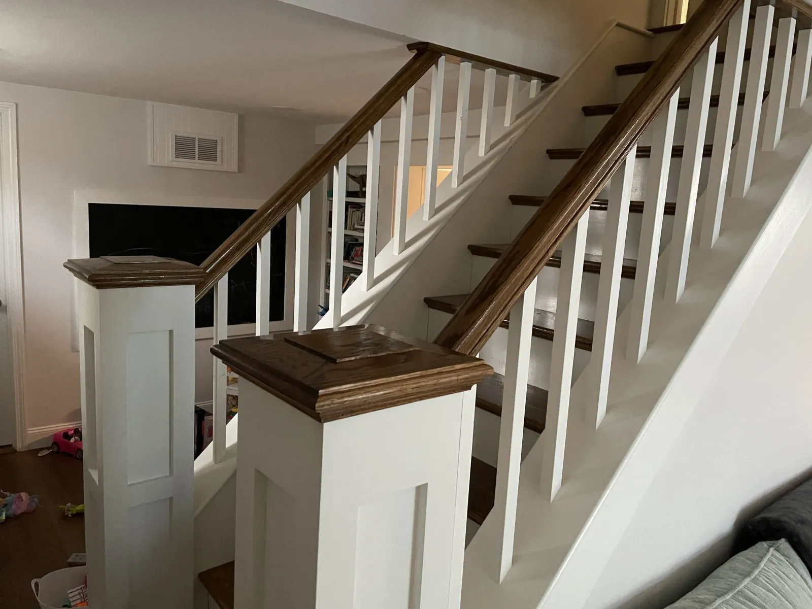 Indoor staircase with wooden handrails and white spindles, leading to an upper level, with a black chalkboard and a bookshelf in the background.
