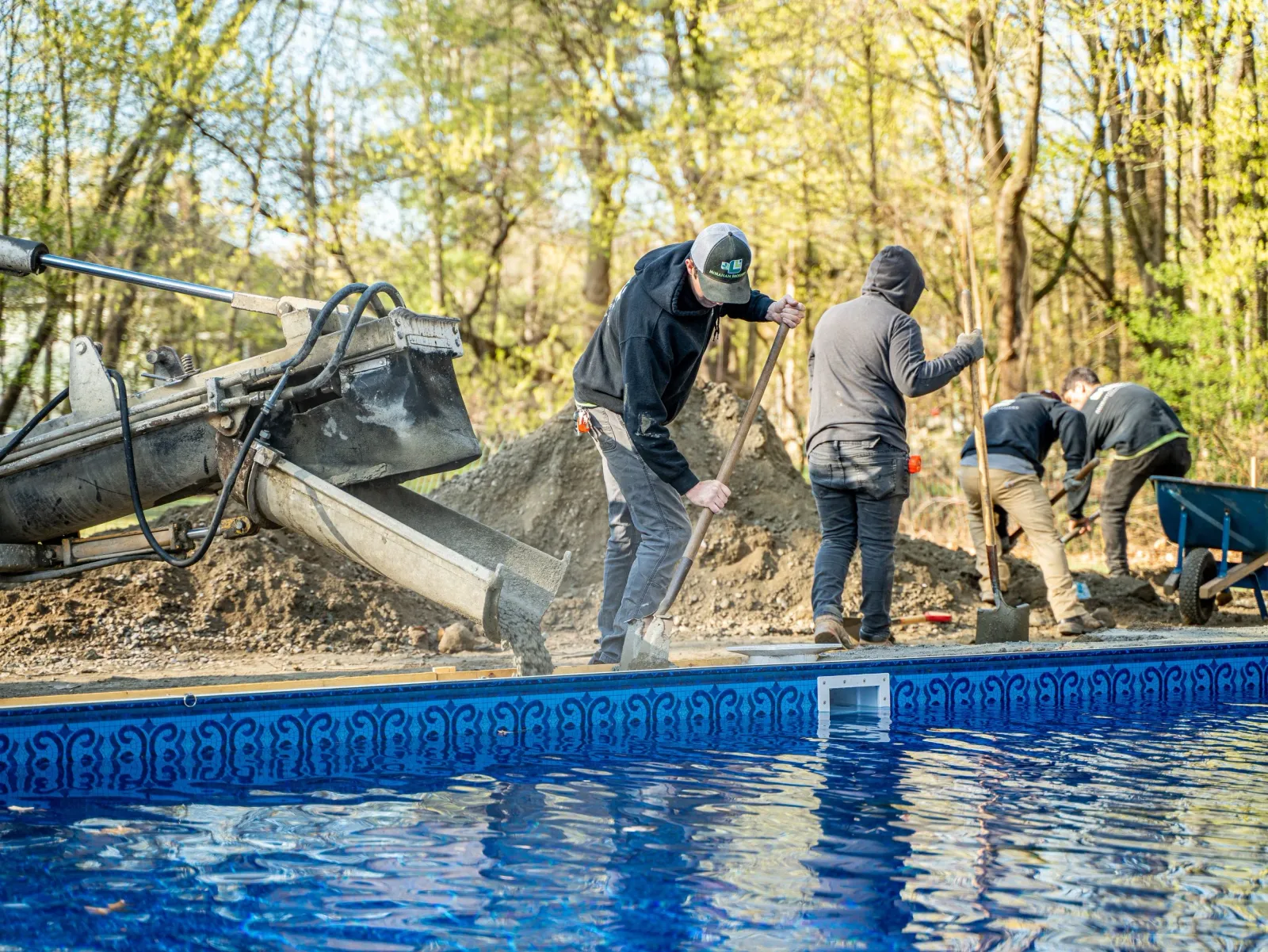 People working on construction site near an in-ground pool, mixing and moving concrete during daytime with trees in background.
