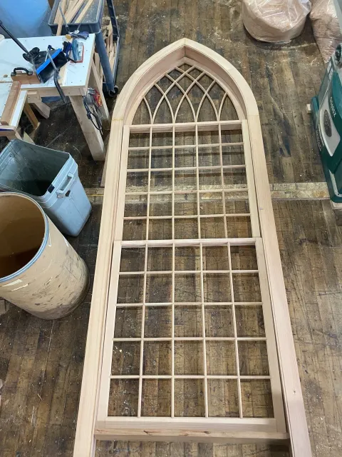 A wooden window frame with a Gothic arch at the top and multiple small glass panes in a workshop setting.