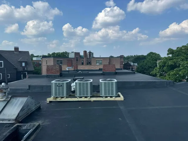 Rooftop with air conditioning units on a wooden pallet; other buildings and blue sky with clouds in the background.