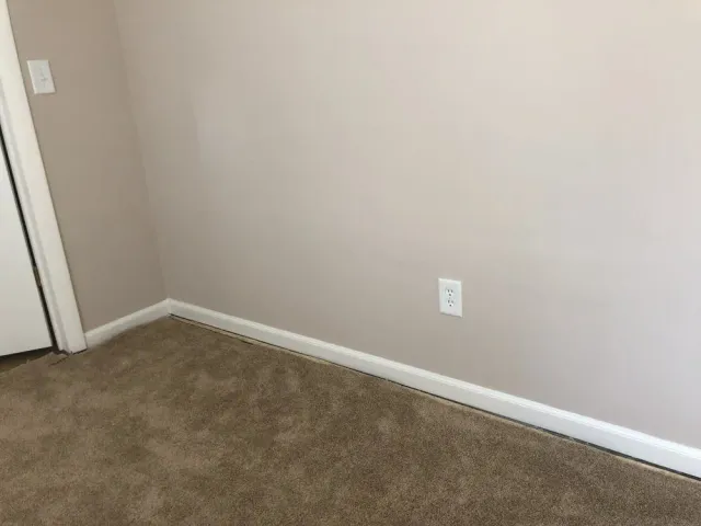 Empty beige room corner with brown carpet, white baseboards, and a door on the left.
