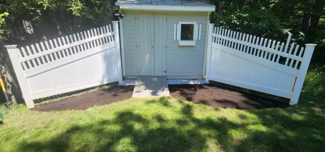 A small gray shed with a white door, flanked by white picket fences on both sides, set in a grassy area.