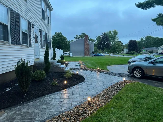 A paved stone walkway illuminated by landscape lights leads to the side entrance of a light-colored house at twilight.