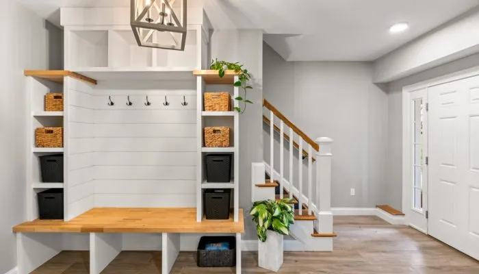 Well-organized entryway with white shiplap-backed built-in unit, natural wood bench top, black coat hooks, woven baskets on open shelves, pendant light overhead, and adjacent staircase with wood treads and white railings.