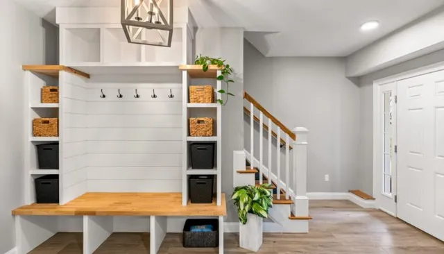 Well-organized entryway with white shiplap-backed built-in unit, natural wood bench top, black coat hooks, woven baskets on open shelves, pendant light overhead, and adjacent staircase with wood treads and white railings.