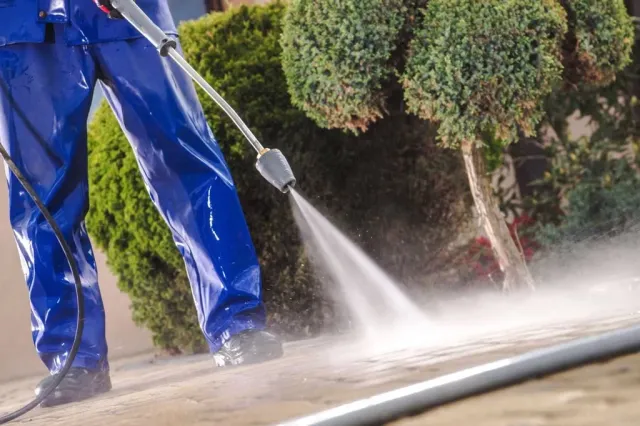 Person in blue waterproof overalls pressure washing a sidewalk near green bushes.