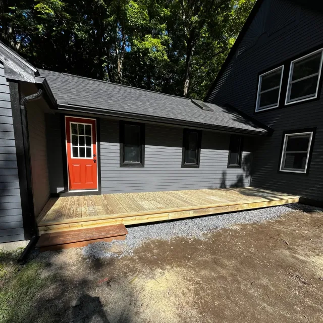 Newly built wooden deck in front of a gray house with a red door and two windows, with a green wooded area in the background.